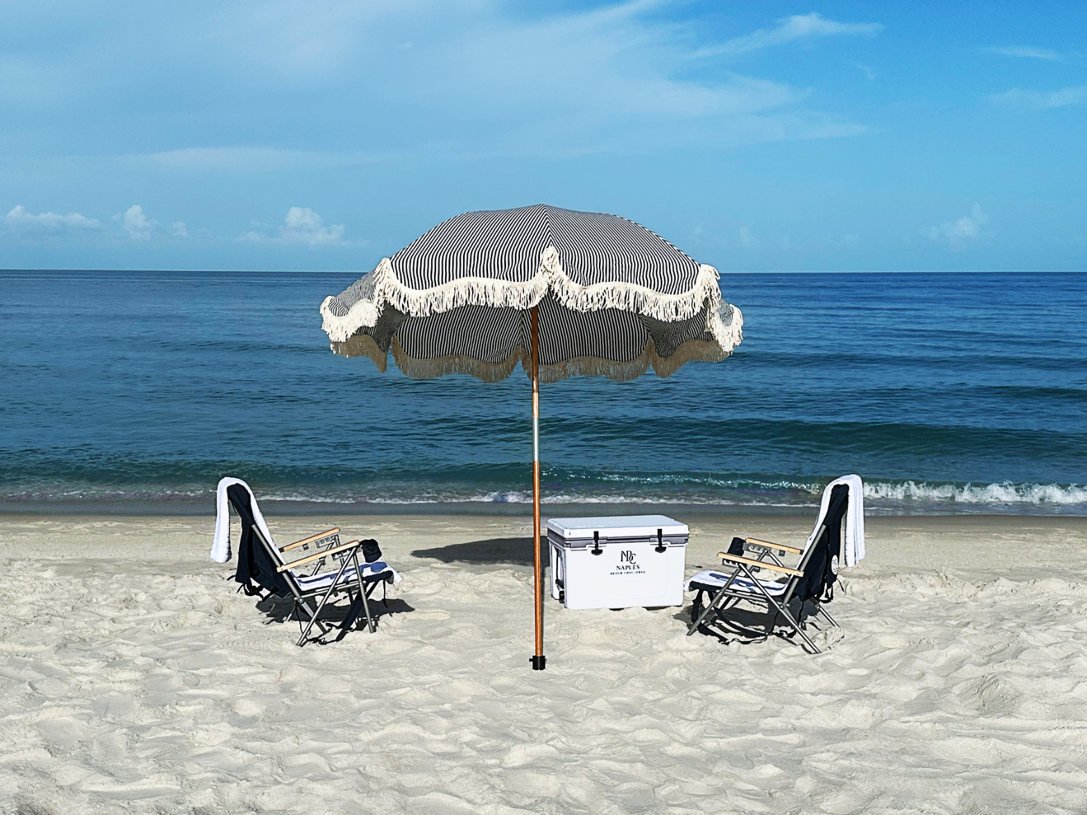 N.B.C. beach setup with striped fringed umbrella, two chairs, and branded cooler on the Naples shoreline