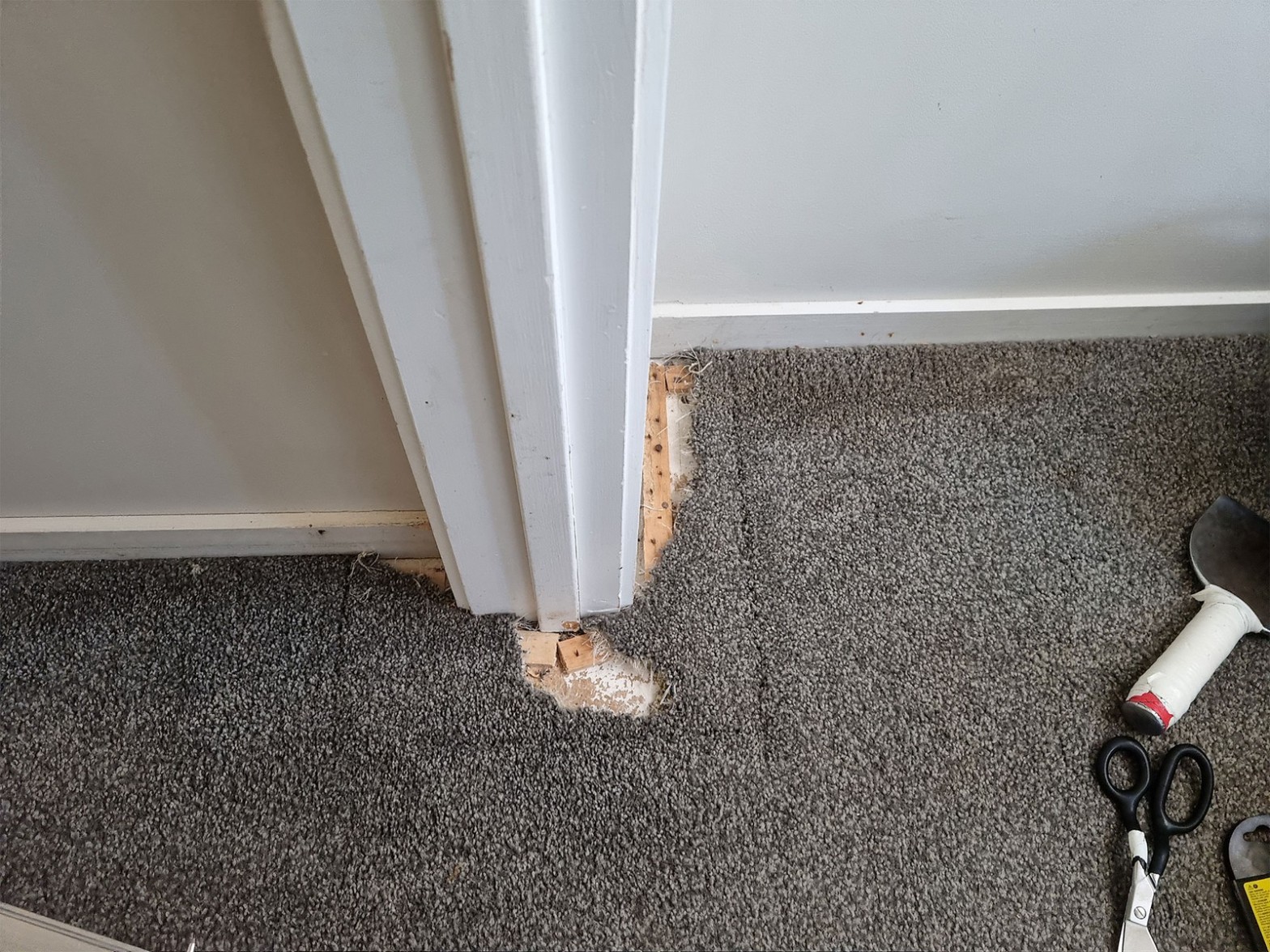 Grey carpet damaged near door frame corner with scissors and repair tools visible