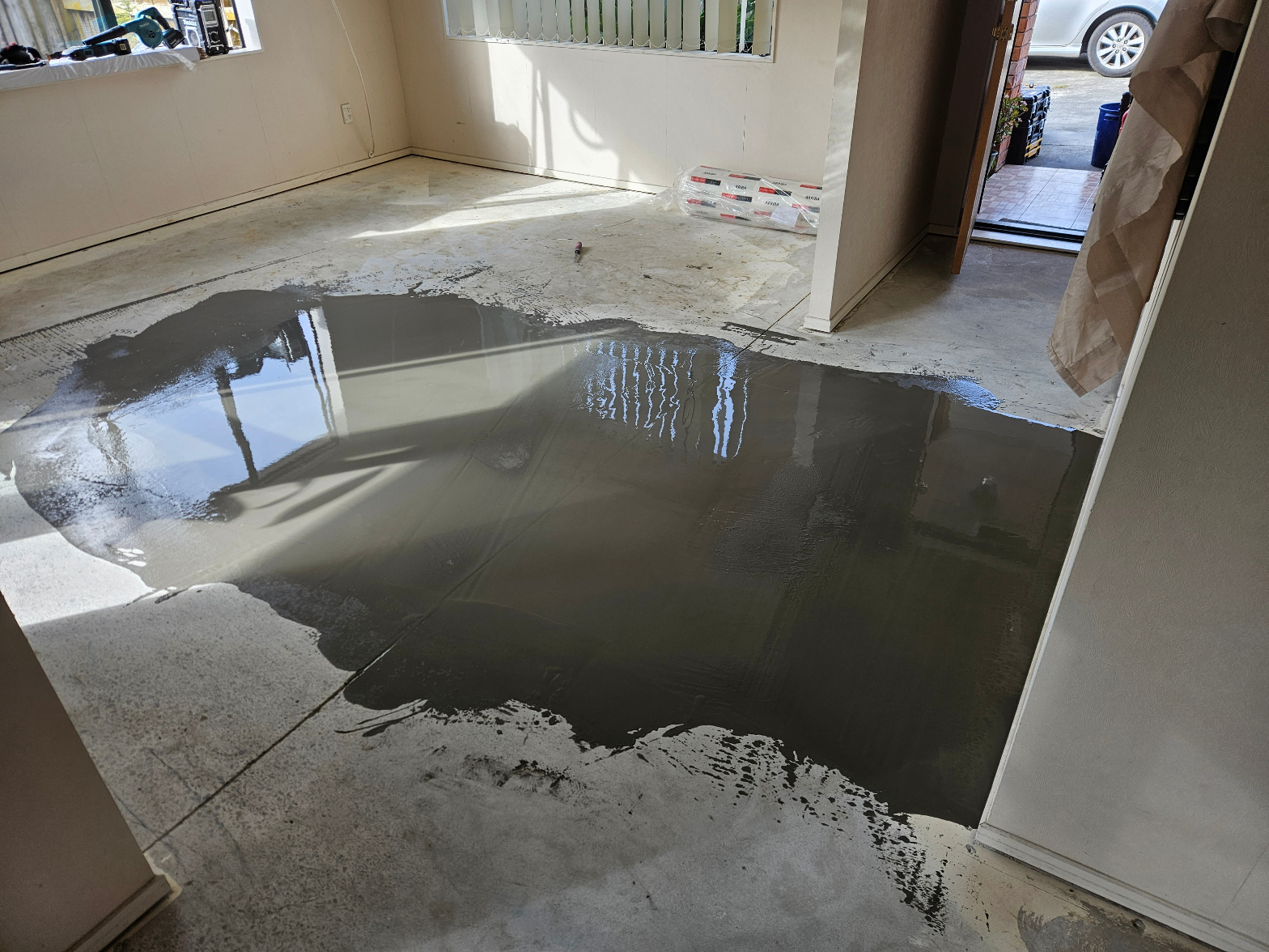 Self-levelling compound freshly poured across a concrete subfloor in an empty room, reflecting light during floor preparation